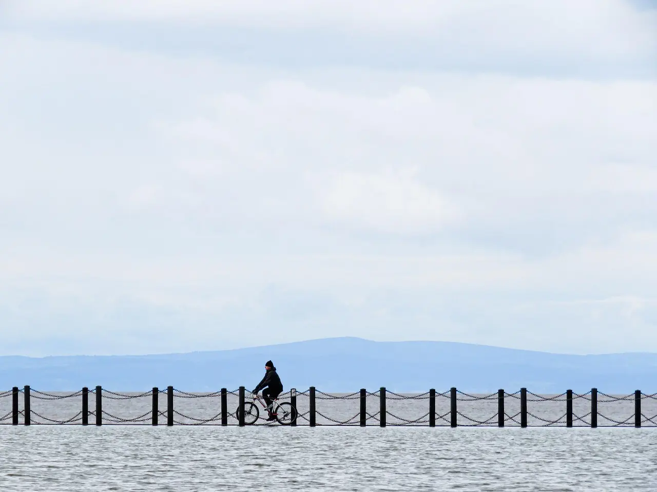 Cyklist på en smal molepromenade over vandet med bjerge i baggrunden